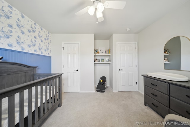 bedroom featuring a nursery area, wainscoting, light carpet, and ceiling fan