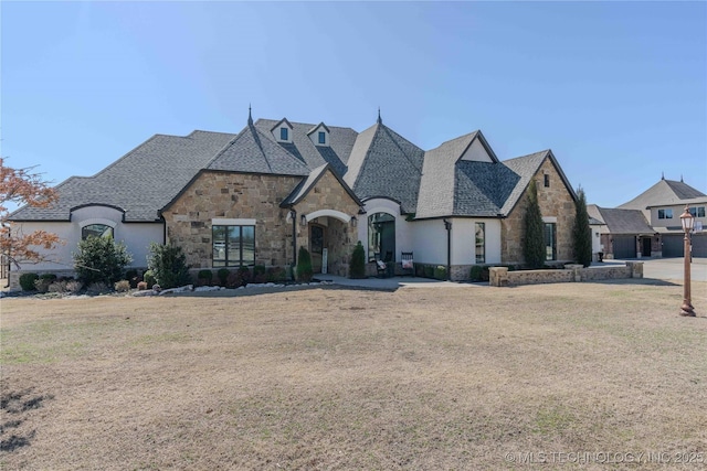 french country style house featuring stone siding, a front lawn, and a shingled roof