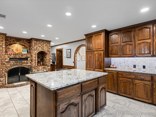 kitchen with a brick fireplace, a center island, light stone counters, crown molding, and decorative backsplash