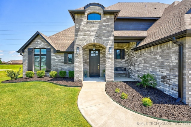 doorway to property featuring brick siding, a lawn, and roof with shingles