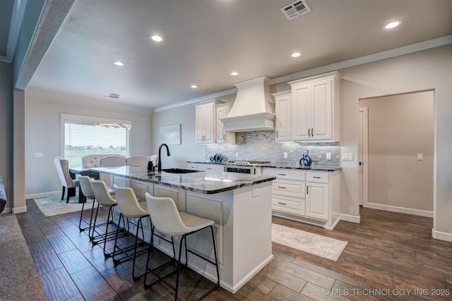 kitchen featuring a kitchen island with sink, custom exhaust hood, visible vents, and white cabinets