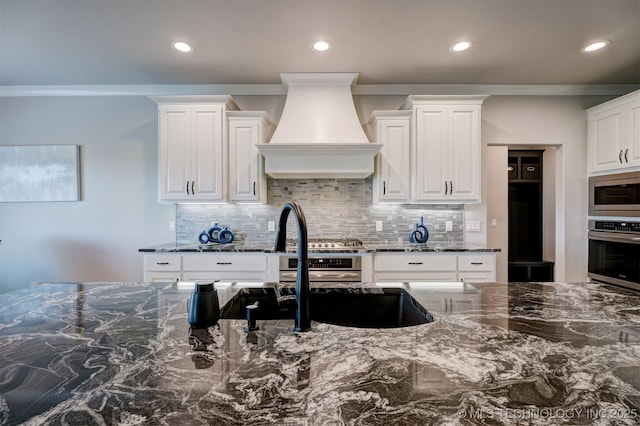 kitchen featuring custom range hood, backsplash, appliances with stainless steel finishes, white cabinetry, and dark stone countertops