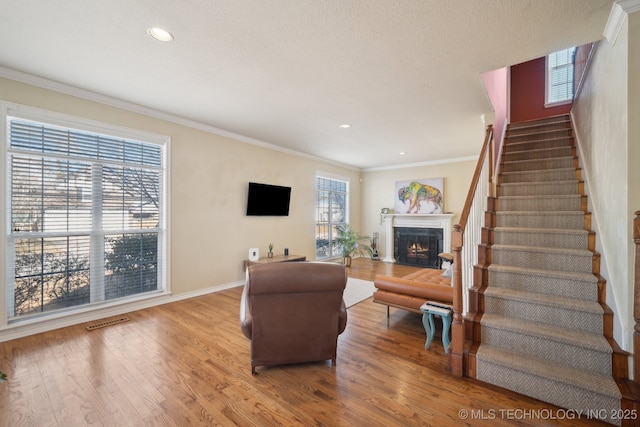 living room featuring a warm lit fireplace, wood finished floors, visible vents, stairs, and ornamental molding