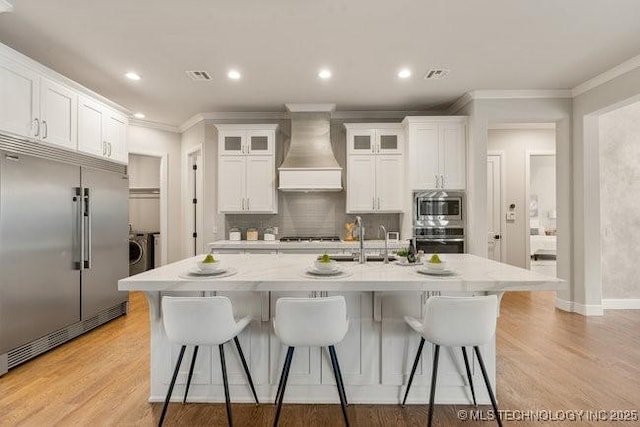 11904 S Toledo Ave, Tulsa, OK 74137 - kitchen featuring white cabinets, wall chimney exhaust hood, glass insert cabinets, built in appliances, and a kitchen island with sink