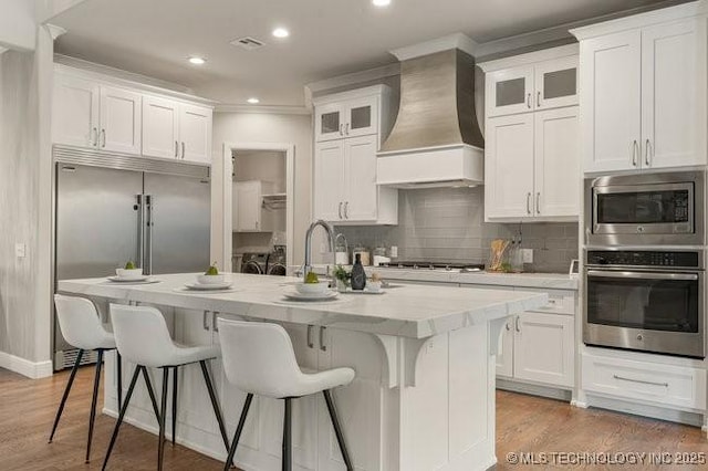 11904 S Toledo Ave, Tulsa, OK 74137 - kitchen featuring built in appliances, a kitchen island with sink, white cabinetry, wall chimney exhaust hood, and glass insert cabinets