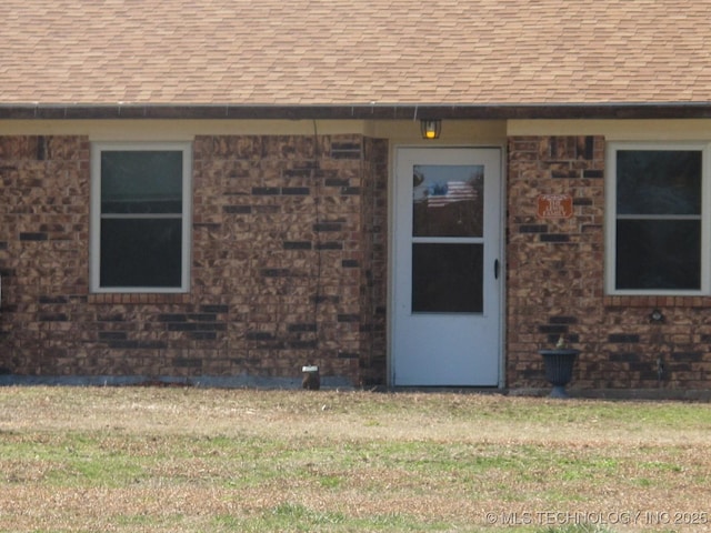 entrance to property featuring a shingled roof, brick siding, and a lawn