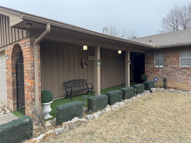 doorway to property featuring a garage, roof with shingles, a yard, board and batten siding, and brick siding