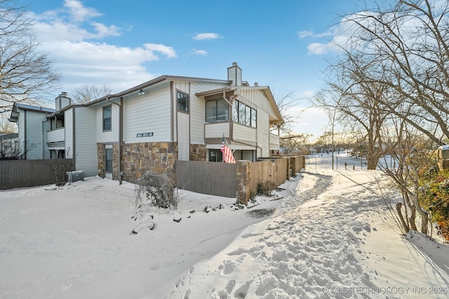 snow covered property with cooling unit, stone siding, fence, and a chimney