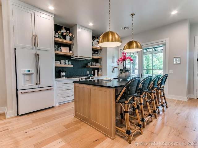 kitchen featuring a center island with sink, light wood finished floors, open shelves, high end white fridge, and decorative backsplash