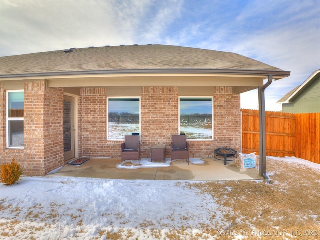 snow covered house featuring an outdoor fire pit, a patio, roof with shingles, fence, and brick siding