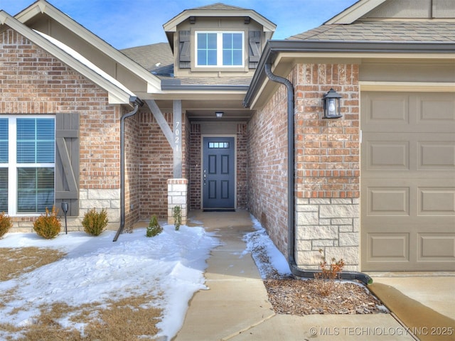 snow covered property entrance with a garage, brick siding, and roof with shingles