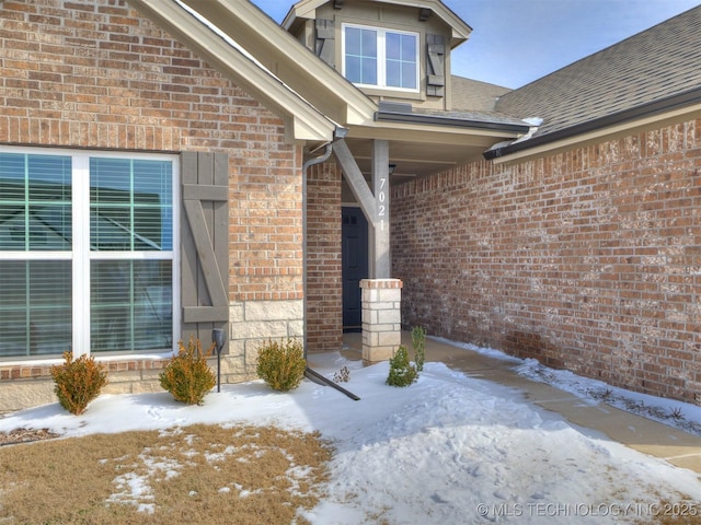 snow covered property entrance with roof with shingles and brick siding