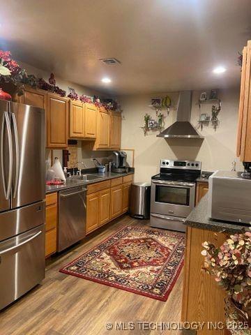 kitchen with stainless steel appliances, a sink, wood finished floors, wall chimney exhaust hood, and dark countertops