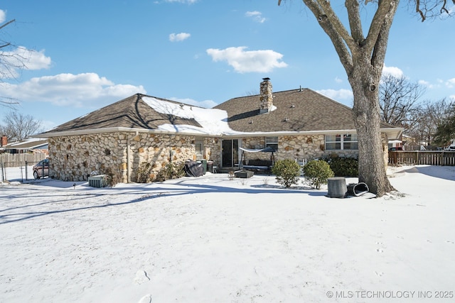 snow covered house featuring stone siding, fence, a chimney, and central air condition unit