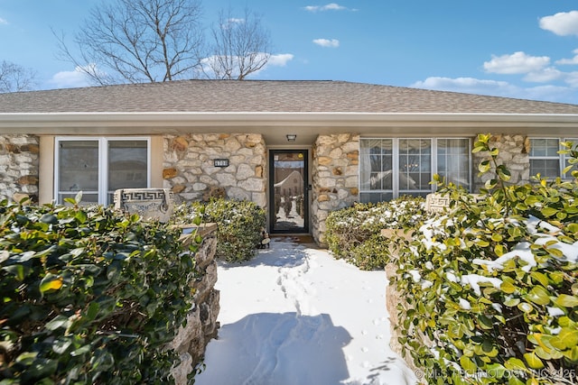 snow covered property entrance featuring stone siding and roof with shingles