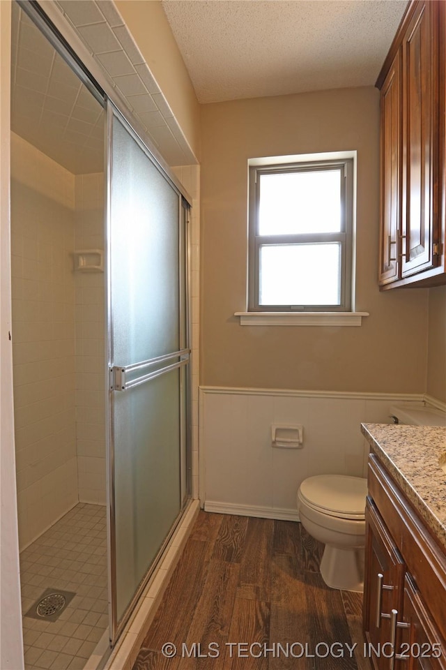 full bathroom with a textured ceiling, toilet, wood finished floors, wainscoting, and a shower stall