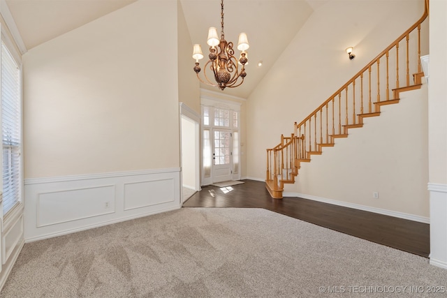 foyer entrance featuring high vaulted ceiling, stairs, wainscoting, dark carpet, and an inviting chandelier