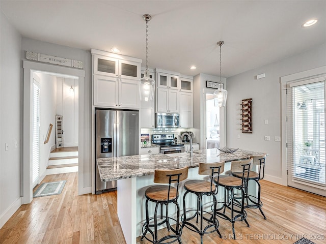 kitchen with light wood-style flooring, white cabinets, appliances with stainless steel finishes, an island with sink, and glass insert cabinets