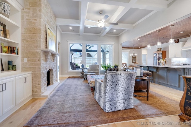 living area with coffered ceiling, a stone fireplace, light wood-style floors, beam ceiling, and ceiling fan with notable chandelier