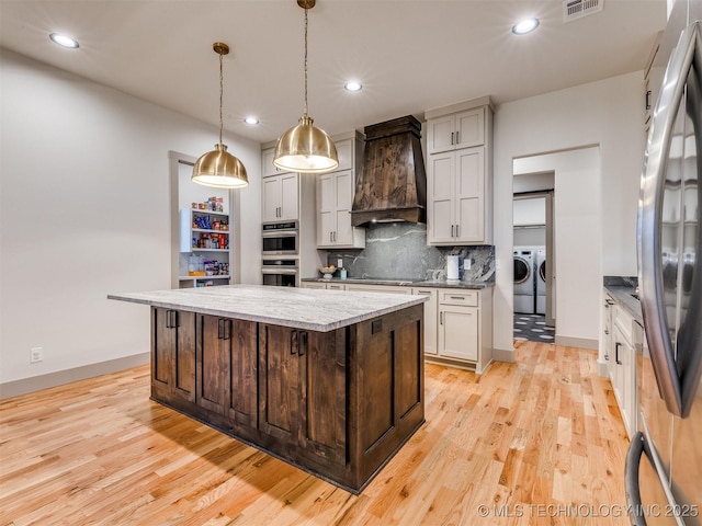 kitchen with stainless steel appliances, a kitchen island, custom exhaust hood, washer and clothes dryer, and pendant lighting