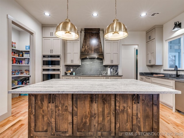 kitchen featuring a kitchen island, stainless steel appliances, light wood-type flooring, premium range hood, and a sink