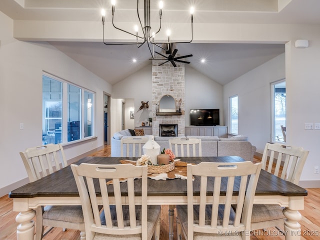 dining area featuring vaulted ceiling with beams, light wood finished floors, a large fireplace, baseboards, and ceiling fan with notable chandelier