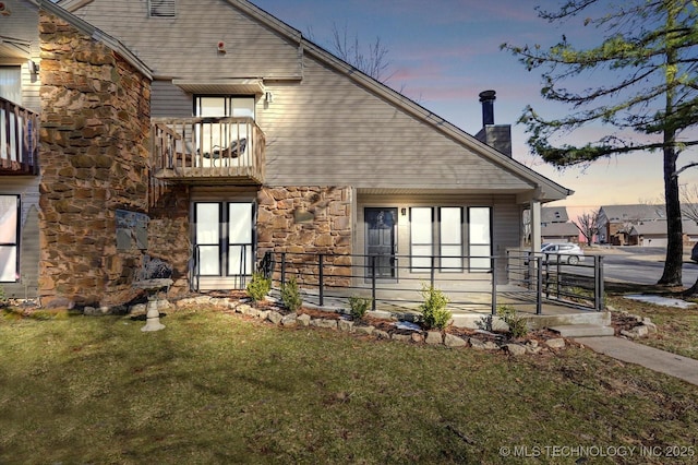 back of house featuring a chimney, stone siding, a lawn, and a balcony