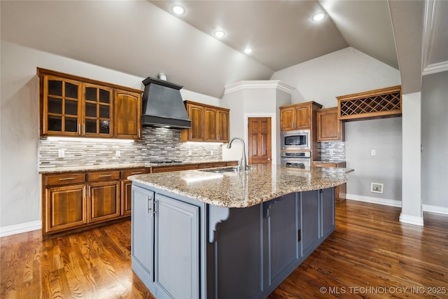 kitchen featuring premium range hood, a sink, appliances with stainless steel finishes, brown cabinetry, and an island with sink