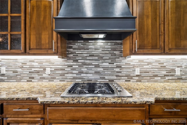 kitchen featuring stainless steel gas stovetop, backsplash, glass insert cabinets, light stone countertops, and premium range hood