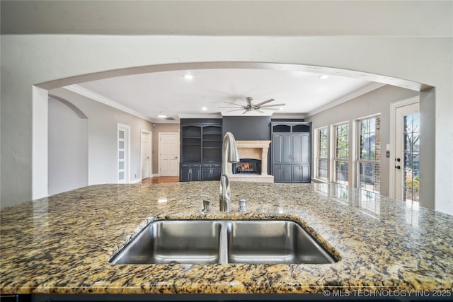 kitchen featuring a warm lit fireplace, stone countertops, arched walkways, ornamental molding, and a sink