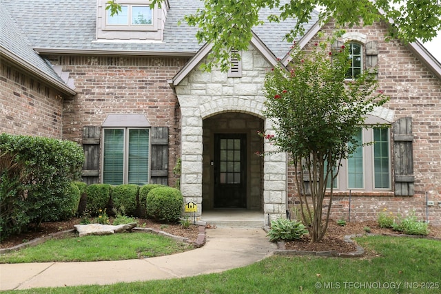 entrance to property featuring a yard, stone siding, brick siding, and roof with shingles