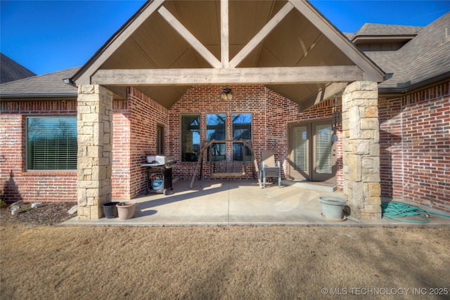 view of exterior entry featuring a patio area, roof with shingles, and brick siding