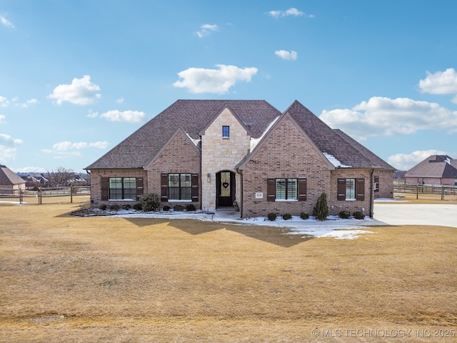 french country inspired facade featuring stone siding, roof with shingles, fence, a front yard, and brick siding