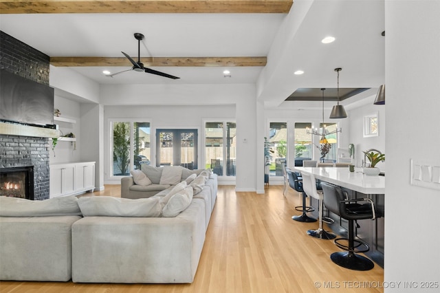 living room featuring a brick fireplace, beamed ceiling, light wood-style flooring, and ceiling fan with notable chandelier