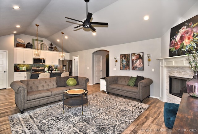 living area featuring arched walkways, dark wood finished floors, a ceiling fan, vaulted ceiling, and a stone fireplace