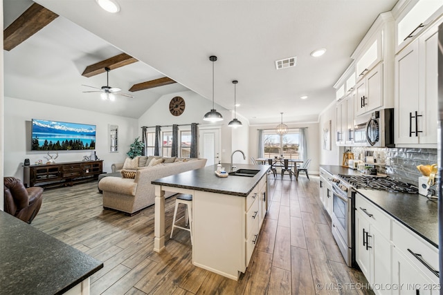 kitchen with a center island with sink, stainless steel appliances, dark countertops, open floor plan, and white cabinetry