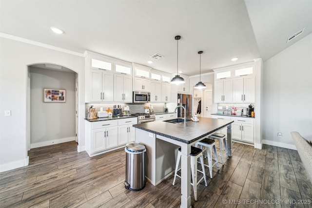 kitchen with glass insert cabinets, stainless steel appliances, white cabinetry, dark countertops, and a center island with sink