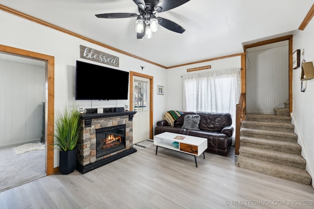 living room featuring ceiling fan, a stone fireplace, ornamental molding, stairway, and light wood finished floors