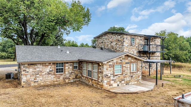 back of house featuring stone siding, a shingled roof, central AC unit, and a balcony