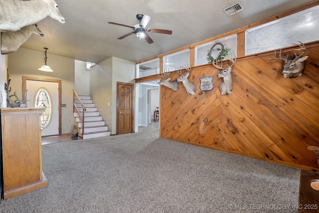 unfurnished living room with carpet floors, visible vents, stairway, a ceiling fan, and wooden walls