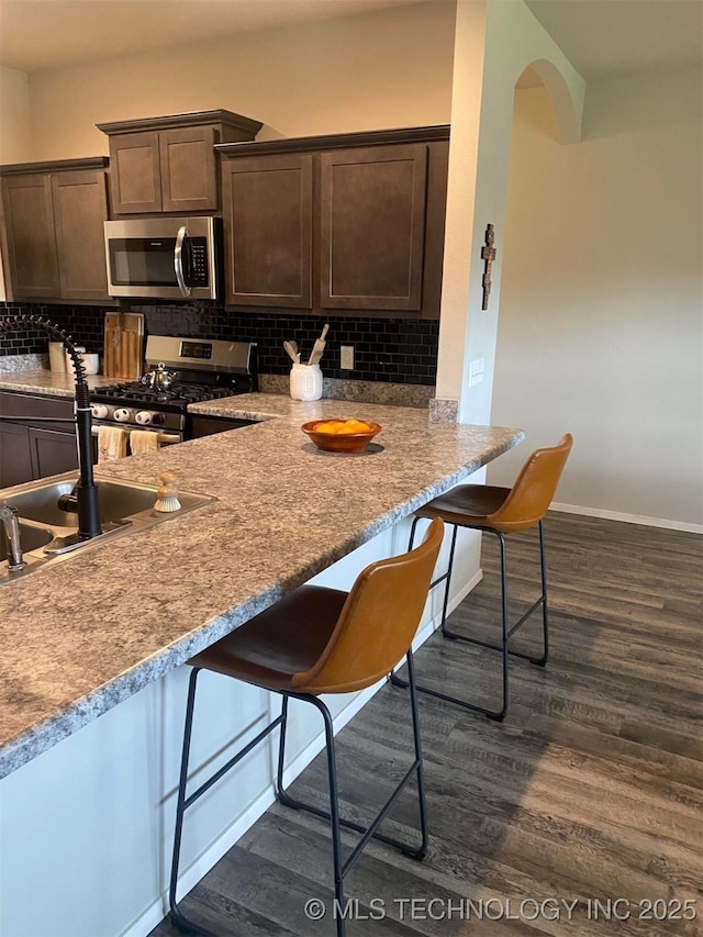 kitchen with light stone counters, a breakfast bar, arched walkways, appliances with stainless steel finishes, and dark brown cabinetry