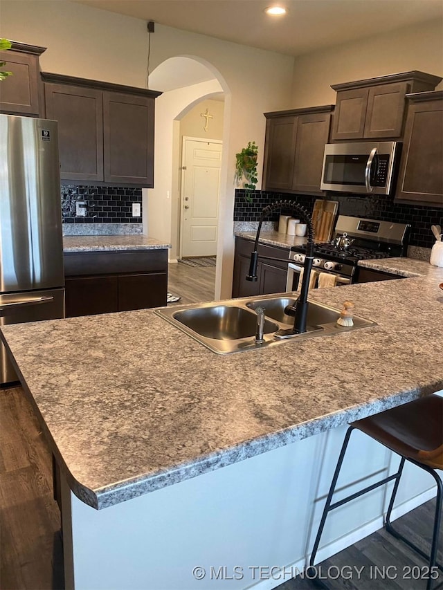 kitchen featuring arched walkways, stainless steel appliances, dark brown cabinets, backsplash, and dark wood-style floors