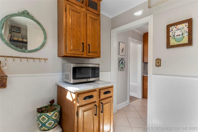 kitchen with tile counters, wainscoting, stainless steel microwave, crown molding, and light tile patterned flooring