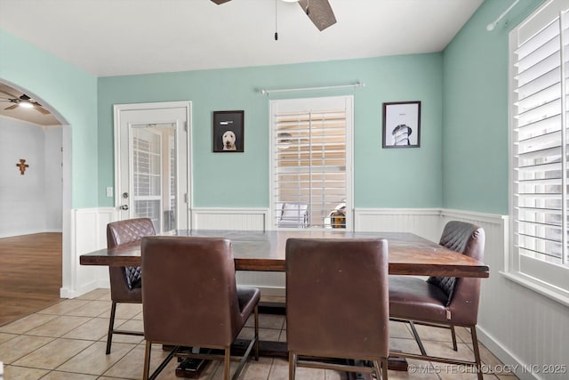 dining area featuring arched walkways, light tile patterned floors, a wainscoted wall, and a ceiling fan