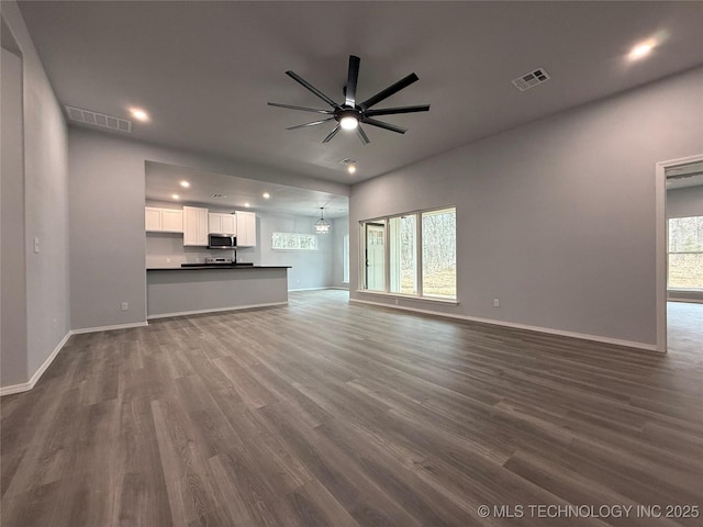 unfurnished living room with a wealth of natural light, visible vents, dark wood-style flooring, and ceiling fan with notable chandelier