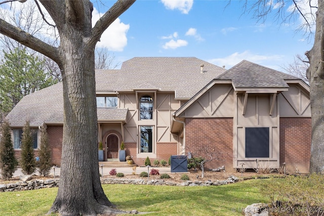 tudor-style house featuring a shingled roof, a front yard, brick siding, and stucco siding