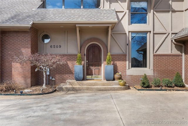 doorway to property featuring stucco siding, a shingled roof, and brick siding