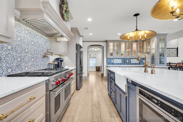 kitchen with light wood finished floors, arched walkways, glass insert cabinets, appliances with stainless steel finishes, and premium range hood