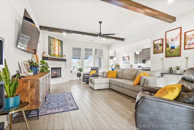 living area featuring a warm lit fireplace, a ceiling fan, light wood finished floors, and beam ceiling