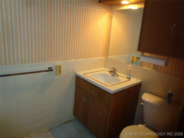 half bath with wallpapered walls, wainscoting, toilet, vanity, and tile walls